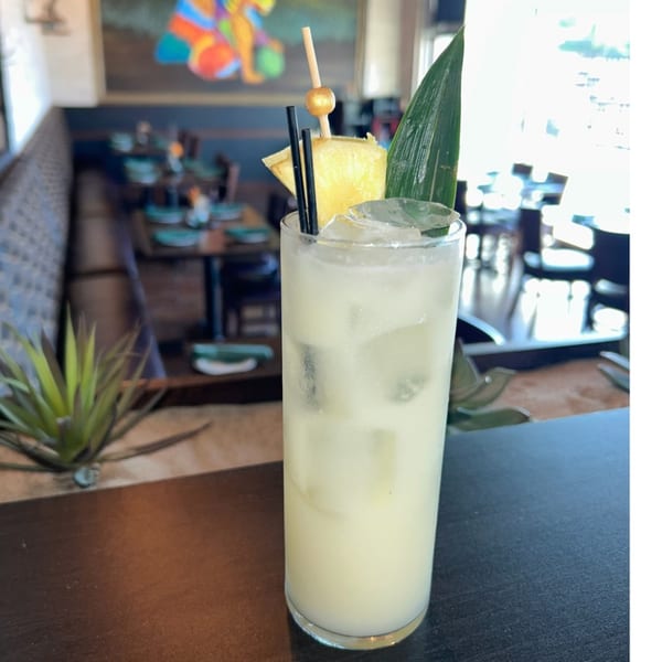 A tall glass of light yellow drink with ice, pineapple, and a green leaf, in a restaurant.