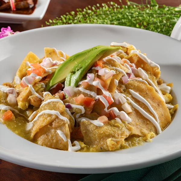 Chilaquiles Verdes with sour cream, diced tomatoes, onions, and avocado slices on a white plate.