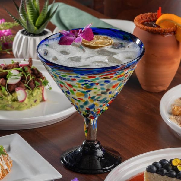 A colorful margarita glass with a drink, lime, and orchid, alongside Mexican dishes.